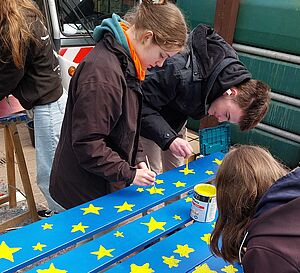 Kinder bemalen Holzbretter mit gelben Sternen auf blauem Grund.